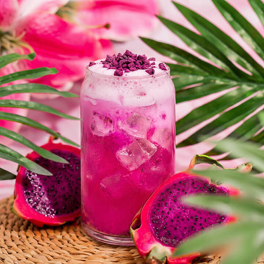 Pink dragon fruit lemonade with ice cubes in a glass, surrounded by dragon fruit and palm leaves.