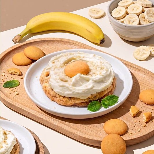 A dessert stack on a white plate, featuring a banana pudding cookie with a wafer cookies on top, garnished with mint leaves. The plate is set on a wooden tray surrounded by freeze dried banana slices, vanilla wafer cookies, and another dessert stack, with a whole banana and a bowl of banana slices in the background.