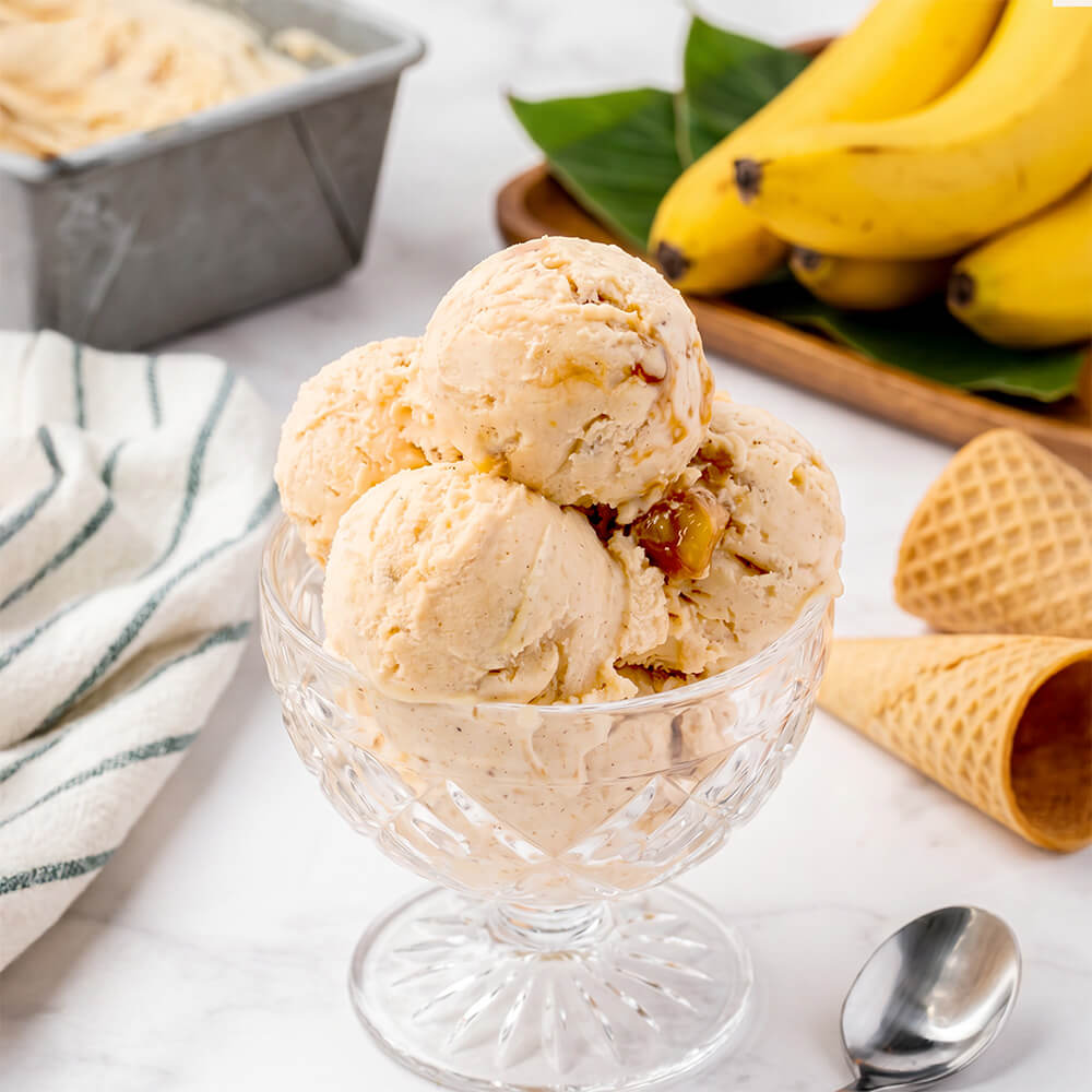 Scoops of banana ice cream in a clear glass sundae bowl, with waffle cones, a spoon, a striped cloth, and bananas in the background.