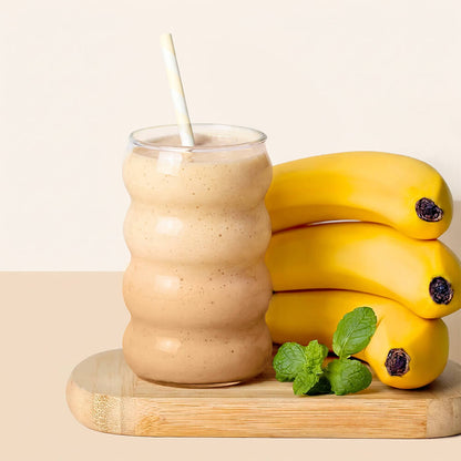 A wavy clear glass filled with a banana smoothie and a white straw, styled on a wooden board with three yellow bananas and mint leaves against a warm beige background.