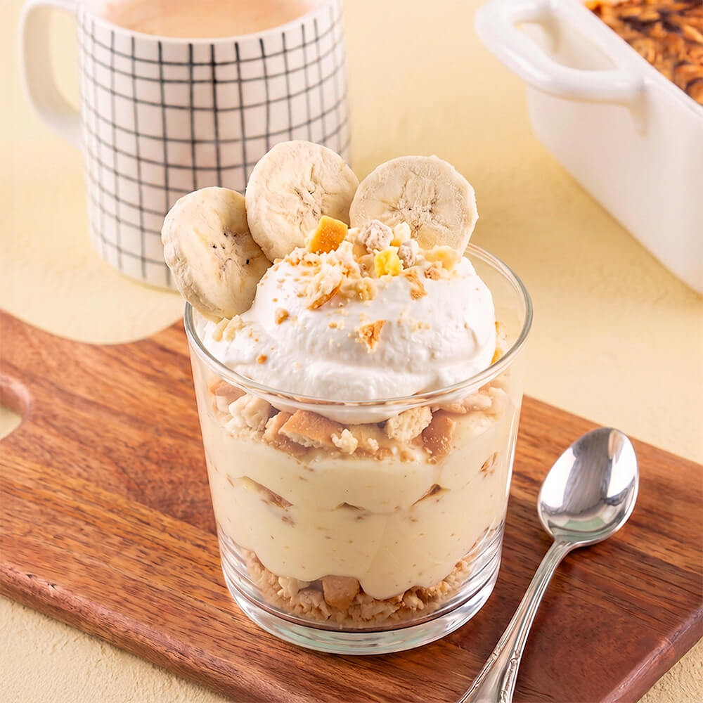 A dessert parfait in a clear glass cup topped with whipped cream, freeze dried banana slices, and crumbled cookie pieces, set on a wooden board with a spoon and a mug in the background.