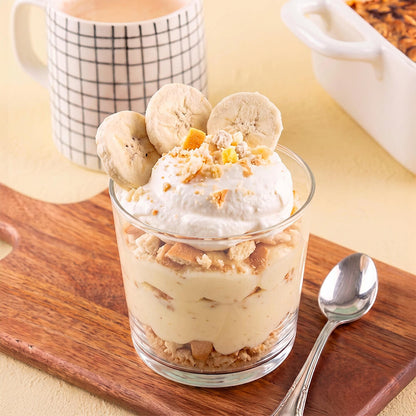 A dessert parfait in a clear glass cup topped with whipped cream, freeze dried banana slices, and crumbled cookie pieces, set on a wooden board with a spoon and a mug in the background.