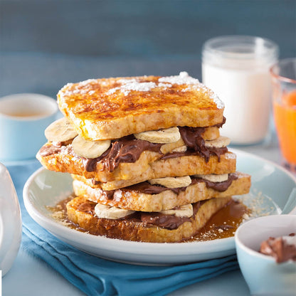 A stack of four French toast slices topped with chocolate spread, freeze dried banana slices, and powdered sugar, served on a white plate with syrup. A small bowl, a cup of coffee, and a glass of orange juice sit in a softly blurred kitchen setting.