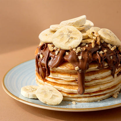 A tall stack of banana pancakes topped with chocolate drizzle, chopped nuts, and freeze dried sliced bananas, served on a light blue plate against a warm brown background.
