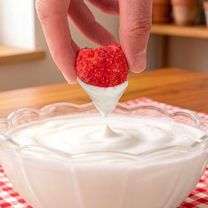Strawberry being dipped into yogurt in a glass bowl on a checkered tablecloth.