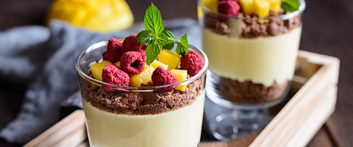 Two dessert cups with layers of cream, fruit, and chocolate crumbles on a wooden tray.