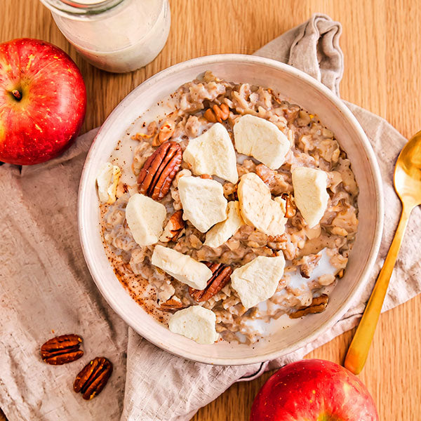 A bowl of oatmeal topped with freeze dried apple slices and pecans, styled on a wooden table with whole apples, a spoon, a napkin, and a glass of milk.