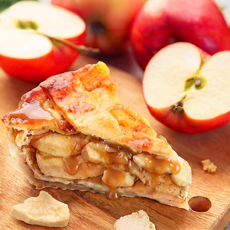 A slice of apple pie with a golden flaky crust, filled with freeze dried sliced apples and drizzled with caramel sauce, presented on a wooden cutting board with whole and halved red apples in the background.