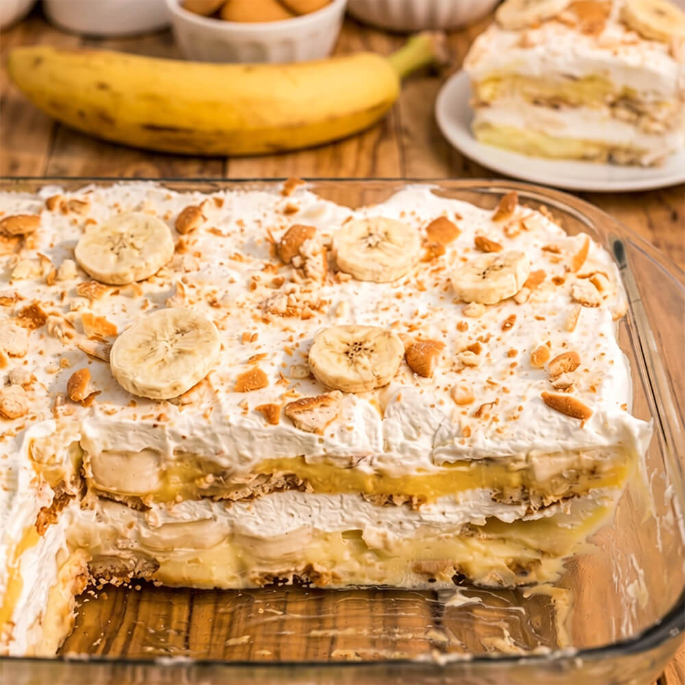 Banana pudding in a glass baking dish with whipped cream, banana slices, and cookie crumbs on top, served on a wooden table with a banana and a slice on a plate in the background.
