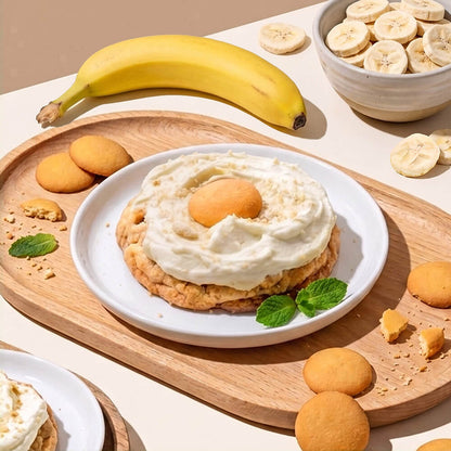 A dessert stack on a white plate, featuring a banana pudding cookie with a wafer cookies on top, garnished with mint leaves. The plate is set on a wooden tray surrounded by freeze dried banana slices, vanilla wafer cookies, and another dessert stack, with a whole banana and a bowl of banana slices in the background.