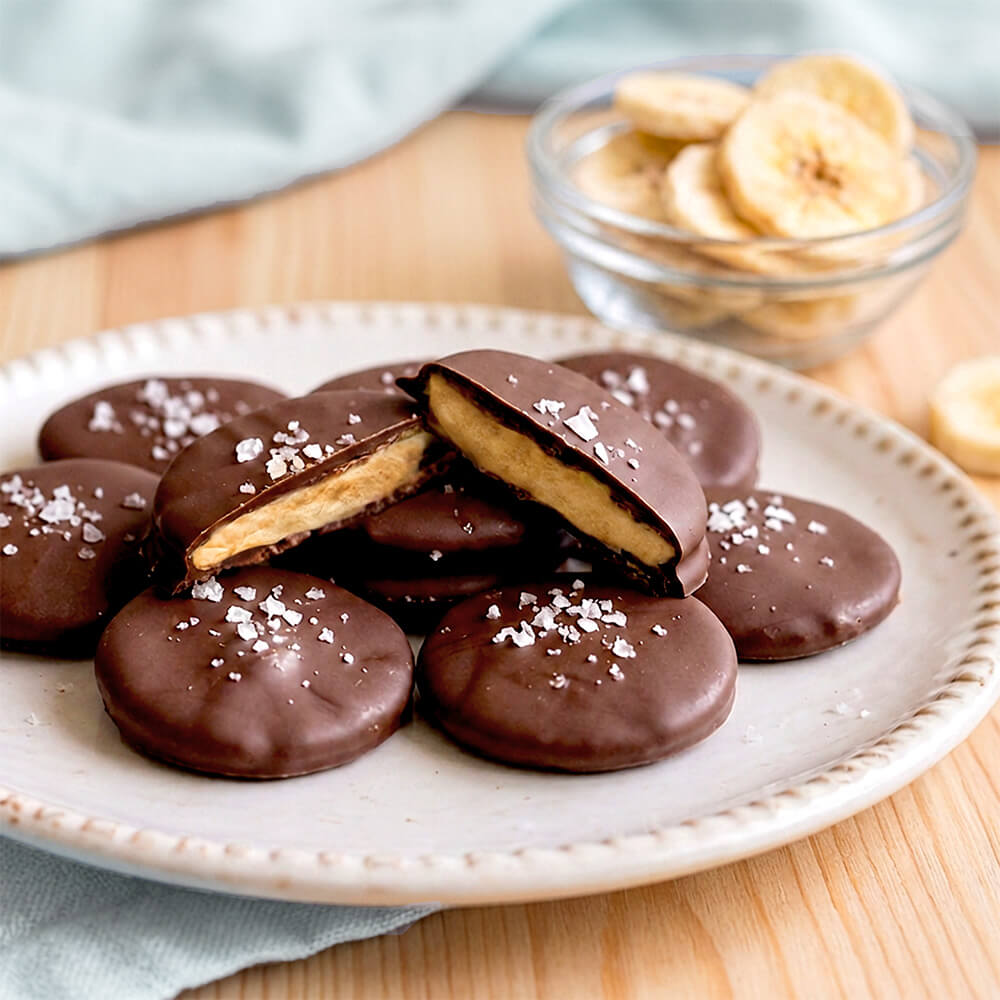 Chocolate covered banana cookies on a white plate, with one cookie broken open to show the banana filling. A small glass bowl of sliced bananas sits in the background on a wooden table.
