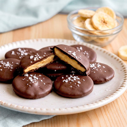 Chocolate covered banana cookies on a white plate, with one cookie broken open to show the banana filling. A small glass bowl of sliced bananas sits in the background on a wooden table.