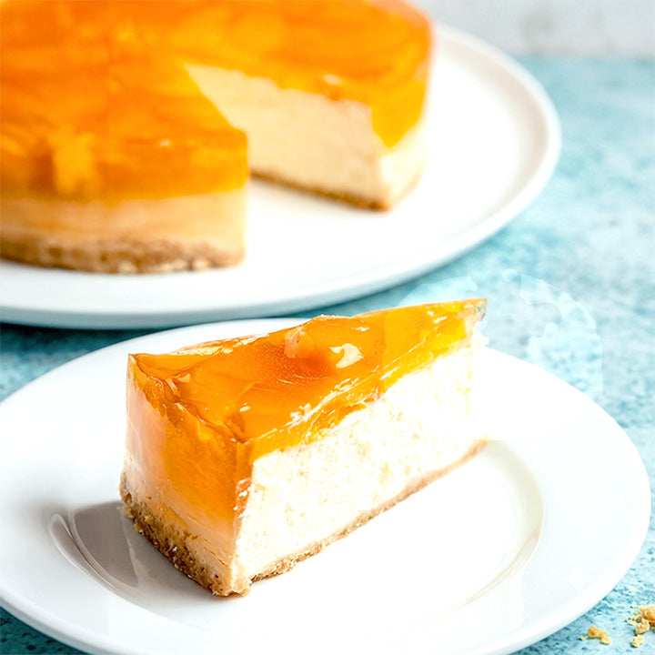 A slice of mango cheesecake with a glossy orange fruit topping sits on a white plate in the foreground, with the full cake on a larger plate blurred in the background.