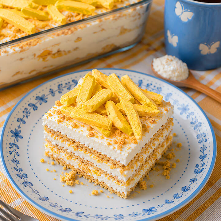 A slice of layered dessert cake with white cream, crumb cookie layers, and freeze dried mango slices on top, served on a blue-and-white patterned plate. A larger dish of the same dessert and a blue cup are visible in the background on a checkered tablecloth.