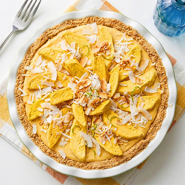 A mango pie with a golden brown crumb crust, creamy yellow filling, freeze dried sliced mango pieces and shredded coconut served in a white pie dish on a striped napkin with a fork and blue bottle in the background.