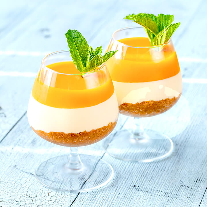 Two layered dessert glasses filled with bright orange mango pudding, creamy white mousse, and a crumb crust, garnished with mint leaves, set on a light blue wooden surface.