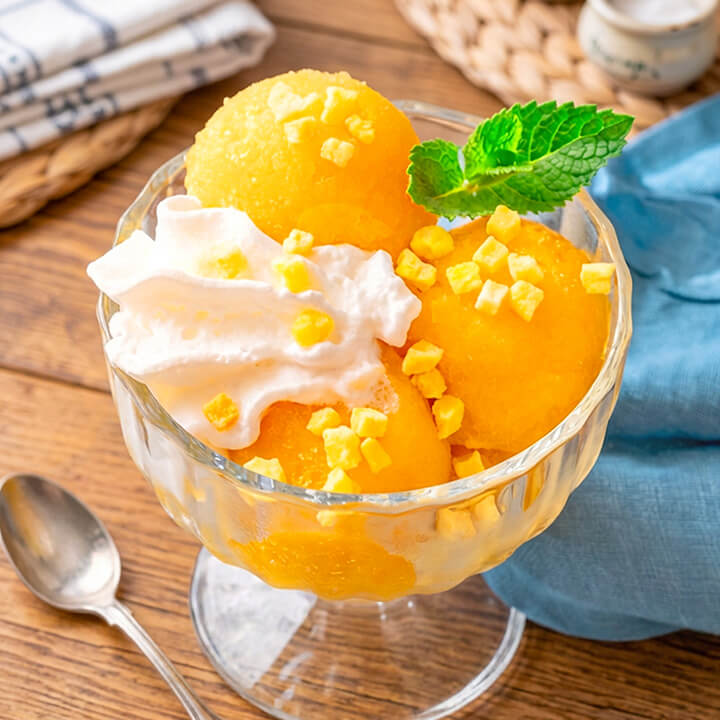 A glass dessert bowl filled with mango sorbet, whipped cream, freeze dried diced mango pieces, and a mint leaf, set on a wooden table with a spoon and blue cloth nearby.